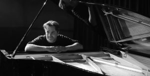 Black and white photo of Darren Pickering sitting behind an open baby grand piano with hands clasped, face turned and looking to the left of focus.